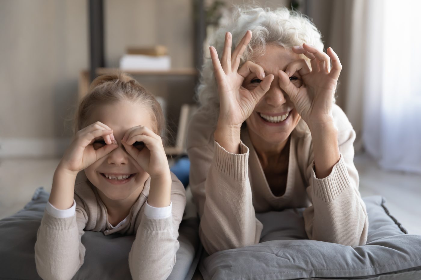 Funny goggles. Portrait of cheerful little girl grandchild lying on floor with senior granny playing having fun look at camera show binoculars glasses of fingers. Good vision and eye care for all ages